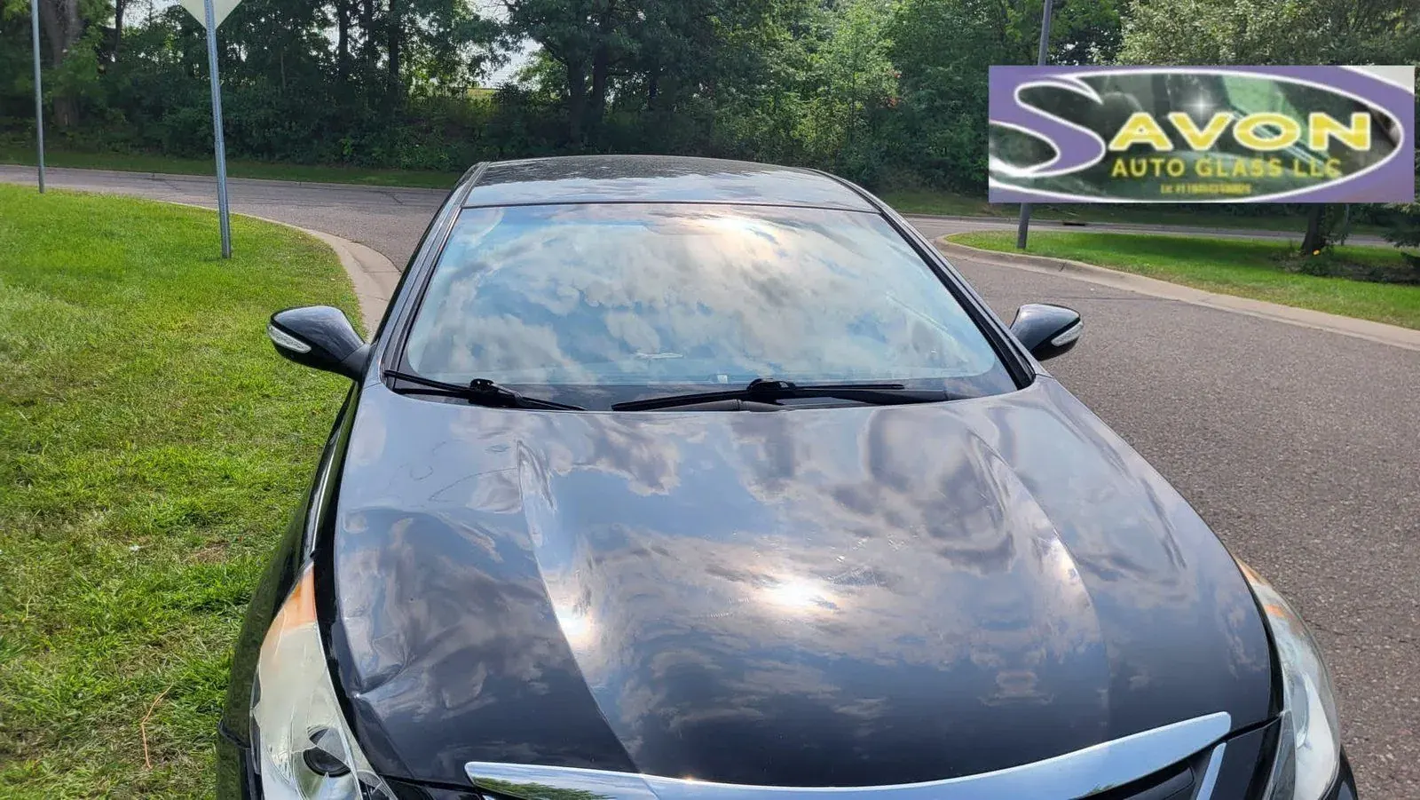 Dark sedan with windshield reflecting sky, parked near Savon Auto Glass business sign